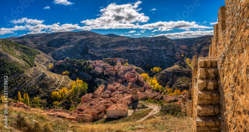 A panoramic photo of Albarracin Spain shot from the wall that was built to protect the ancient city with color of autumn on the surrounding mountains and a bright blue sky with white clouds above.