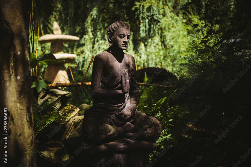 Stone sitting Buddha statue illuminated by the sun in shadow of trees in the japanese garden during the sunny summer day with peacfully zen atmosphere