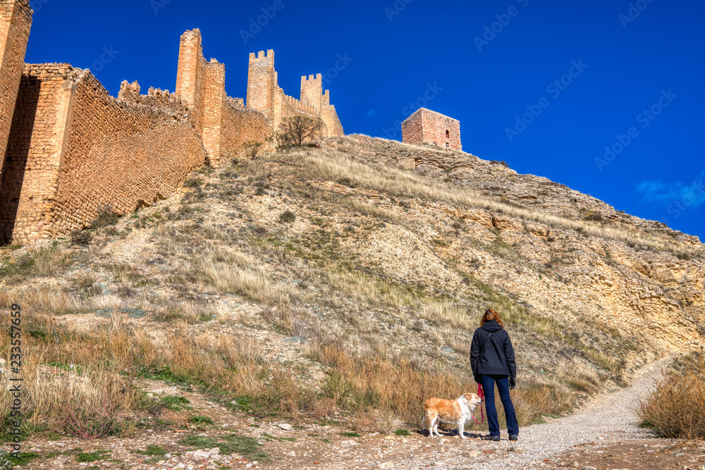 A horizontal photo of a woman in a dark jacket holding the leash of a blonde border collie looking up at the ancient stone wall of Albarracin Spain on a hiking path and a deep blue sky above.