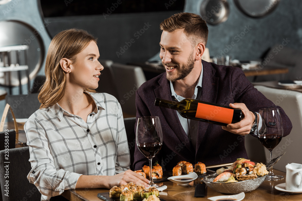 Happy young adult couple having dinner while man pouring wine in glasses in restaurant Happy young adult couple having dinner while man pouring wine in glasses in restaurant