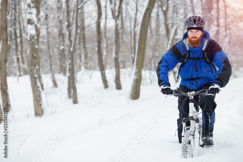 Focused young man with long beard riding a mountain bike through deep powder snow in a forest. Active sport lifestyle in cold weather.