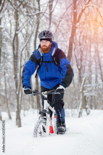 Fit young man with long beard riding a mountain bike through deep powder snow in a forest. Active sport lifestyle in cold weather.