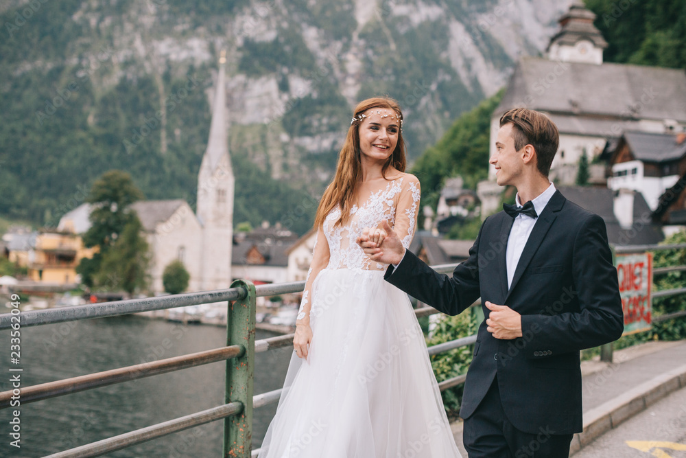 Fototapeta premium A beautiful wedding couple walks on a background old cathedral, lake and mountains in a fairy Austrian town, Hallstatt.