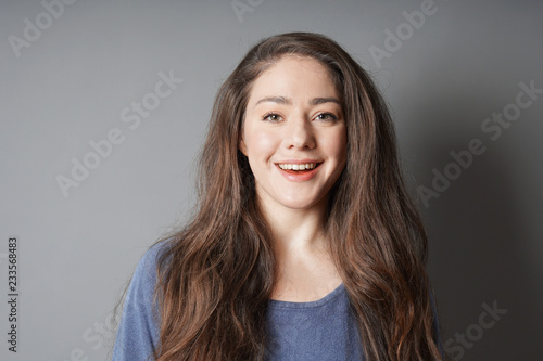 happy young woman in her 20s with natural make-up and brunette long hair smiling toothy smile - against gray background with copy space