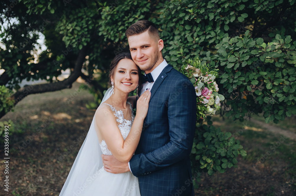 Beautiful newlyweds hugging and smiling in a green garden with trees. Young bride and cute bride with a bouquet are standing in the park. Wedding photography. Stylish portrait. Beautiful newlyweds hugging and smiling in a green garden with trees. Young bride and cute bride with a bouquet are standing in the park. Wedding photography. Stylish portrait.