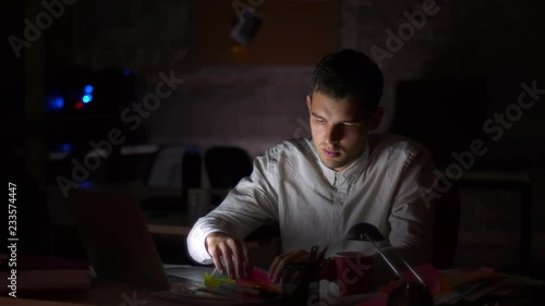 Shooting of dark office, light from screen ar shining on his face while he is sitting looking at computer and checking papers, holding them with focused confident face, indoor illustration photo