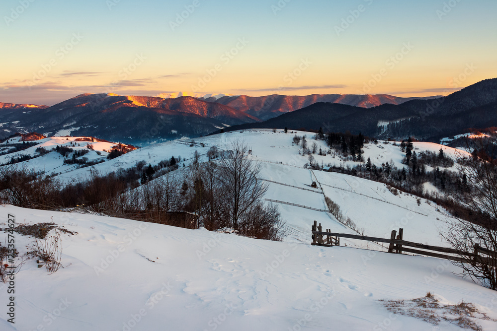 winter countryside of Carpathian mountains. beautiful landscape at sunrise. rural fields on a hill covered in snow. snowy peak of distant ridge shine on the sun light. valley in blue shade