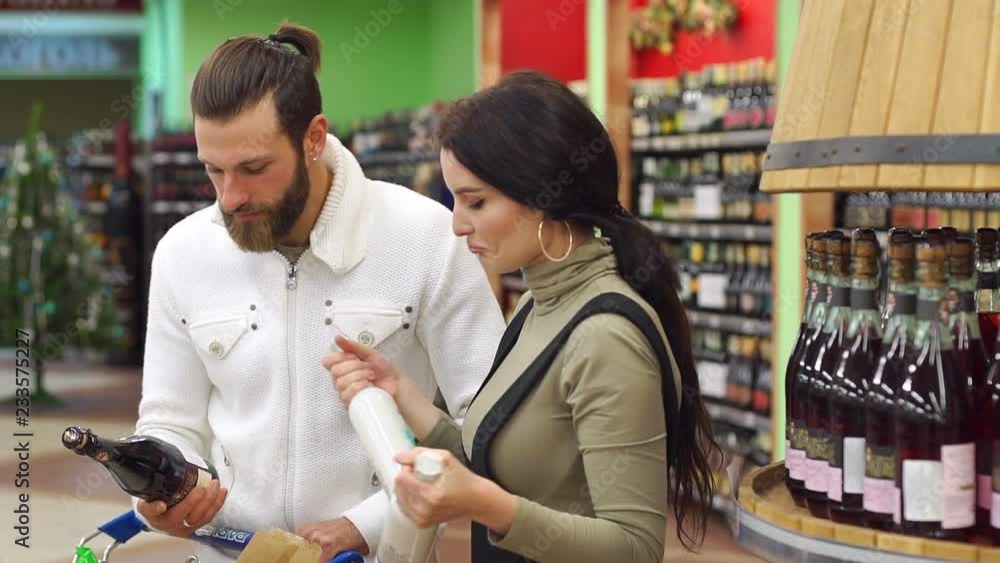 Choosing wine for dinner. Happy fashionable couple choosing wine together while standing in wine store. Beautiful couple is smiling while choosing alcoholic beverage in the supermarket. Slow motion. Choosing wine for dinner. Happy fashionable couple choosing wine together while standing in wine store. Beautiful couple is smiling while choosing alcoholic beverage in the supermarket. Slow motion.