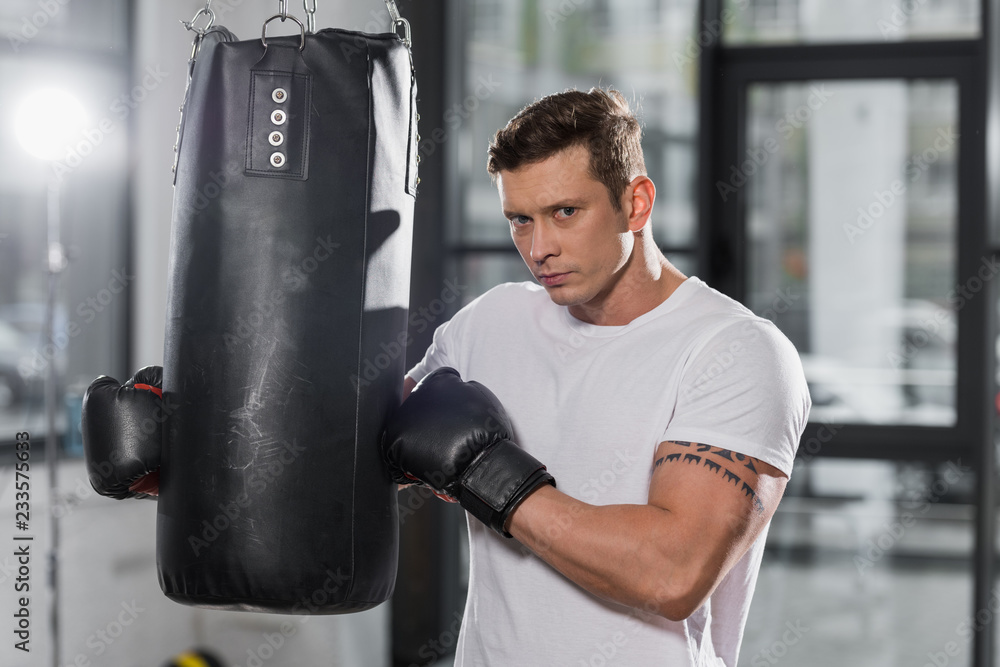 handsome muscular boxer holding punching bag and looking at camera in gym