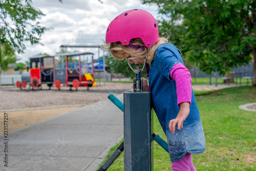 Young girl in pink outfit and pink helmet riding a kick-scooter in the park having a drink from fountain