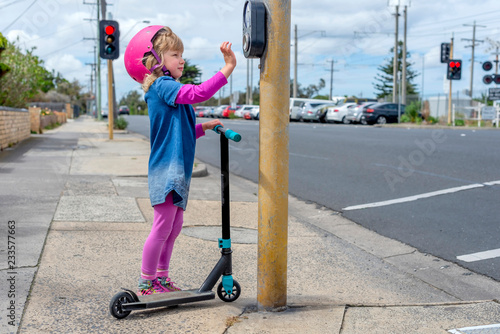 Young girl in pink outfit and pink helmet riding a kick-scooter pressing button at pedestriam crossing