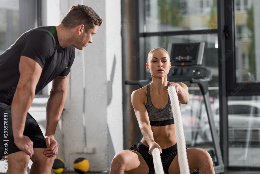 attractive athletic bodybuilder working out with ropes in gym, trainer looking at her attractive athletic bodybuilder working out with ropes in gym, trainer looking at her