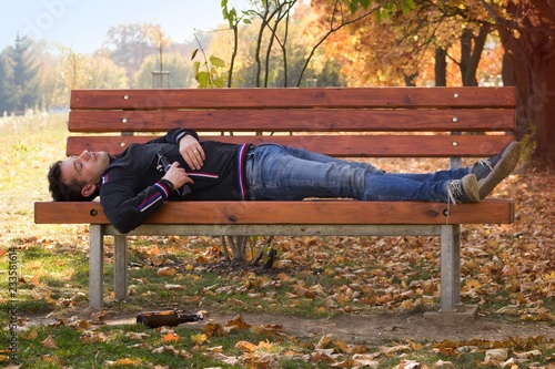 A drunk young man sleeping on a bench in the day.A young man holding a bottle of alcohol in her hand and sleeping on a bench. The problem of alcoholism among young people.