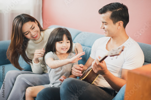 Happy Cheerful smiling Young Family mother father and daughter sitting on the blue sofa, father and daughter play a guitar, rose pink color background wall