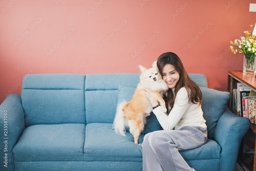 Cheerful young woman holding her puppy with black nose. Indoor portrait of smiling girl with dark long hair posing with dog on rose pink color background and blue sofa at home.
