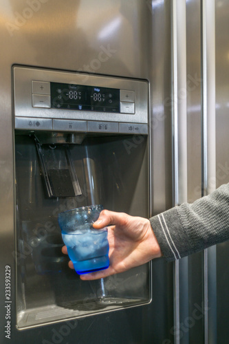 Male hand is pouring cold water and ice cubes from dispenser of home fridge. photo