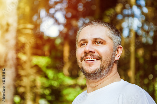 Portrait of a smiling surprised man with beard standing and looking ahead against a green trees in sunny forest