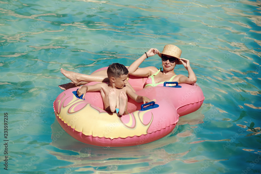 Mother and son enjoying their holidays in aqua park. They are swimming in a rubber ring along the lazy river, smiling and having fun.