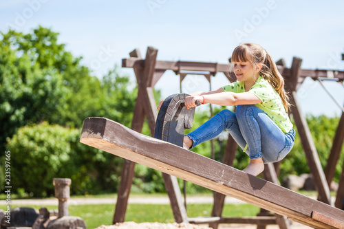 Portrait of little smiling girl child playing outdoor in park in children playground at sunny day in spring. Lifestyle natural shot with blurred background. Weatherproof, wooden outdoor playground