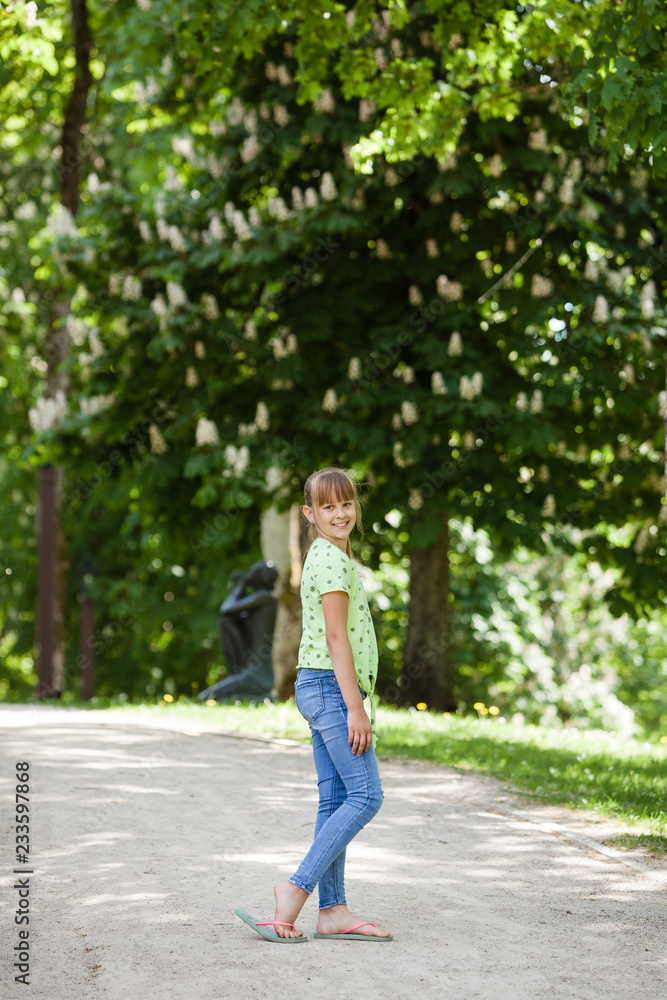 Portrait of little smiling girl child walking outdoor in park at sunny day in spring. Blooming chestnut tree in the background. Teen is wearing jeans and green shirt. Lifestyle shot in nature. 