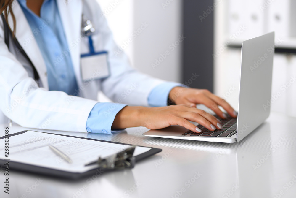 Unknown doctor woman typing on laptop computer while sitting at the desk in hospital office. close-up of hands. Physician at work. Medicine and health care concept Unknown doctor woman typing on laptop computer while sitting at the desk in hospital office. close-up of hands. Physician at work. Medicine and health care concept