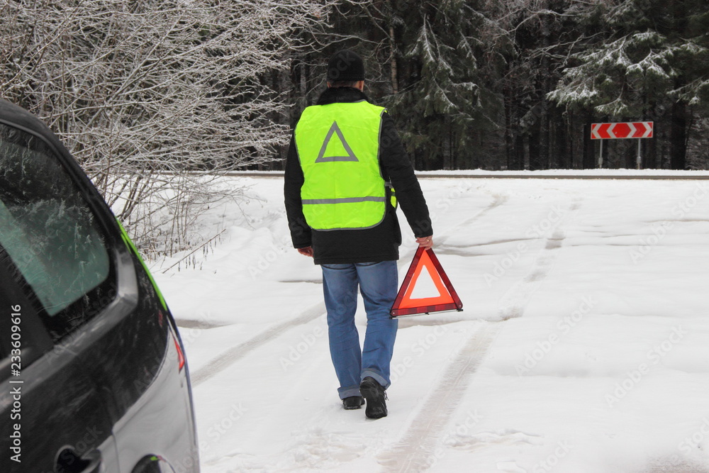 Vehicle safety at a stop, a traffic accident - a man driver in a black jacket and a bright yellow fluorescent signal vest goes to put up an emergency stop sign - red triangle on a forest snow road