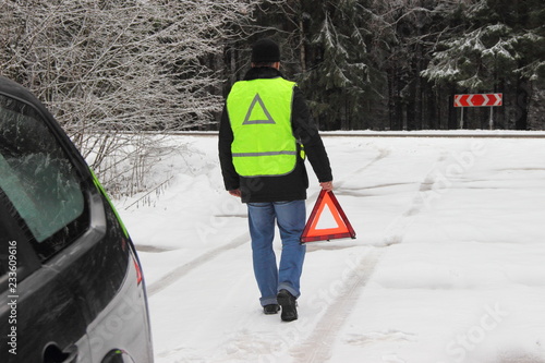 Obraz na płótnie Vehicle safety at a stop, a traffic accident - a man driver in a black jacket and a bright yellow fluorescent signal vest goes to put up an emergency stop sign - red triangle on a forest snow road w salonie