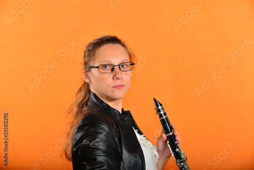 A girl with glasses in a black jacket holds a clarinet in her hands and looks into the camera, on a yellow background.