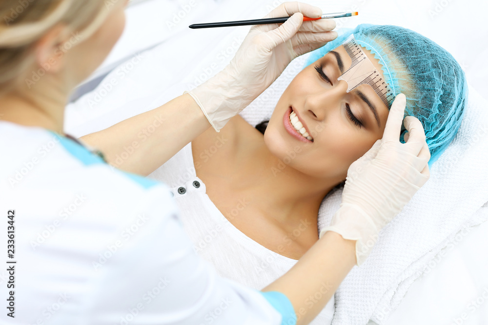 Naklejka premium Professional beautician is going to correct eyebrow line at woman face using cosmetic ruler. Permanent brow makeup in beauty salon, closeup. Cosmetology treatment