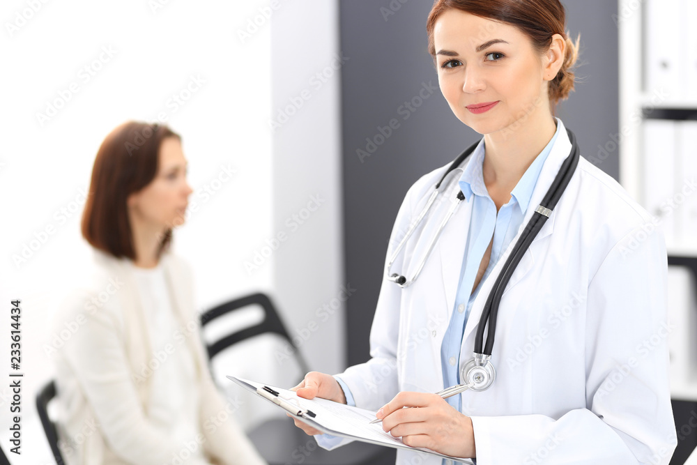 Doctor woman at work. Portrait of female physician filling up medical form while standing near reception desk at clinic or emergency hospital. Patient woman sitting at the background. Medicine concept Doctor woman at work. Portrait of female physician filling up medical form while standing near reception desk at clinic or emergency hospital. Patient woman sitting at the background. Medicine concept