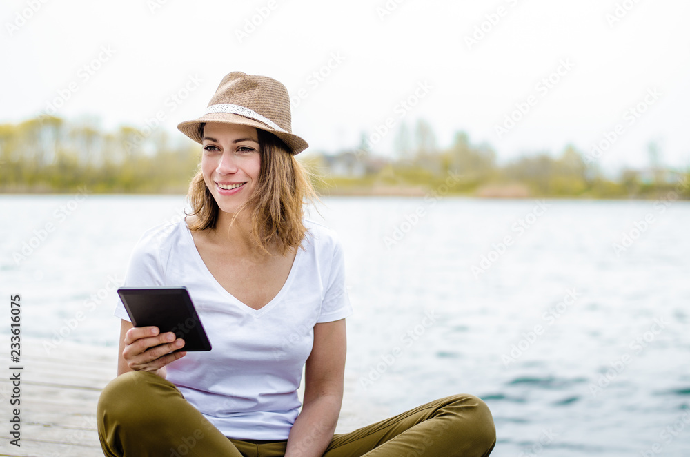 Pretty young woman holding e-book and smiling by the lake.