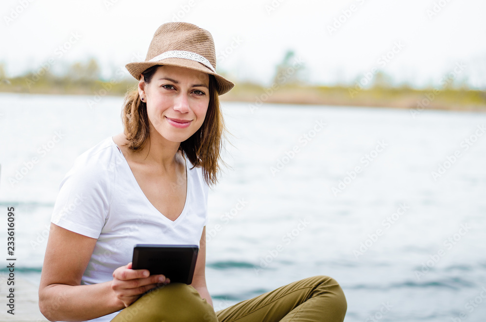 Girl by the lake holding e-book, smiling and looking at camera