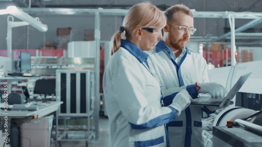 On High Tech Factory Female and Male Engineers Talk and Use Computer for Programing Electronic Machinery for Printed Circuit Board Surface Mount Assembly Line. Production of PCB with SMT Machinery. On High Tech Factory Female and Male Engineers Talk and Use Computer for Programing Electronic Machinery for Printed Circuit Board Surface Mount Assembly Line. Production of PCB with SMT Machinery.