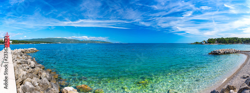 Beautiful summer panoramic coastline seascape. Big long pebble beach in harbor at calm crystal clear azure turquoise water. People rest on the beach. Unrecognizable blurred faces person.