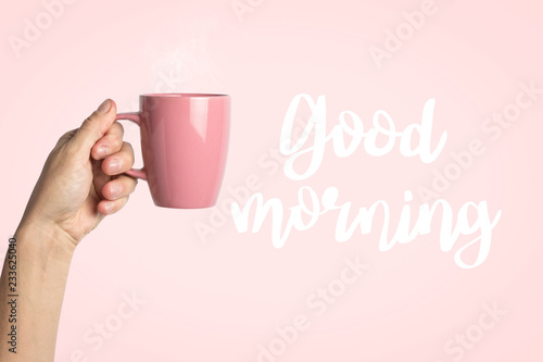 Female hand in clothes holding a purple cup with hot coffee or tea on a pink background. Added text Good morning. Breakfast concept with hot coffee or tea