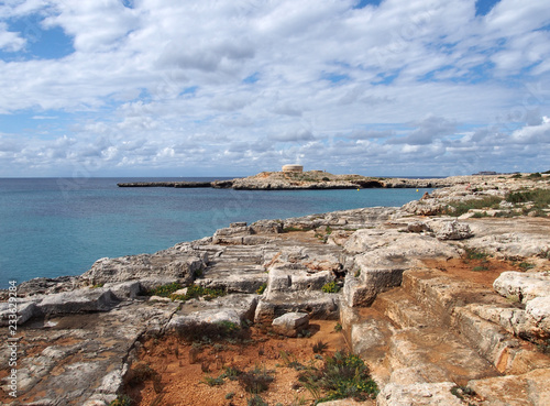 a scenic view of the bay at cala santandria in menorca with carved cliffs and old fort in the distance with blue summer sky photo