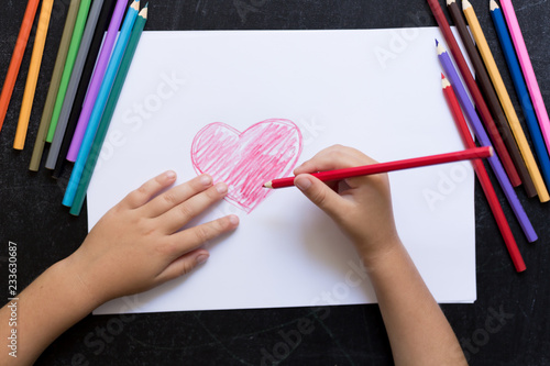 Kid's hands with pencil draws heart on white paper. Mother's day concept. Celebration. Hand made card. Top view.