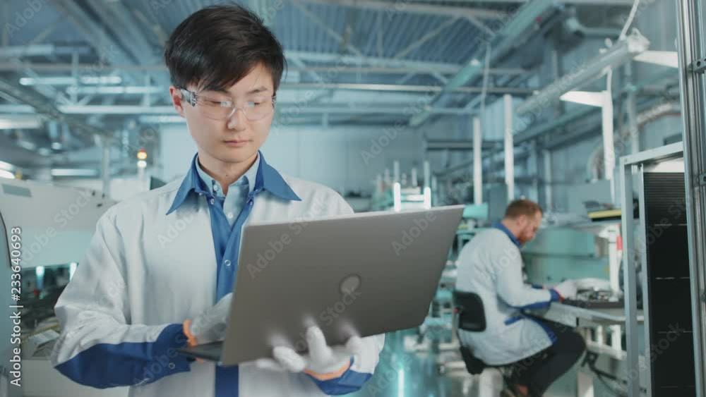 On High-Tech Factory Asian Engineer Uses Laptop Computer for Programing Pick and Place Electronic Machinery for Printed Circuit Board Surface Mount Assembly Line. Production of PCB with SMT Machinery. On High-Tech Factory Asian Engineer Uses Laptop Computer for Programing Pick and Place Electronic Machinery for Printed Circuit Board Surface Mount Assembly Line. Production of PCB with SMT Machinery.