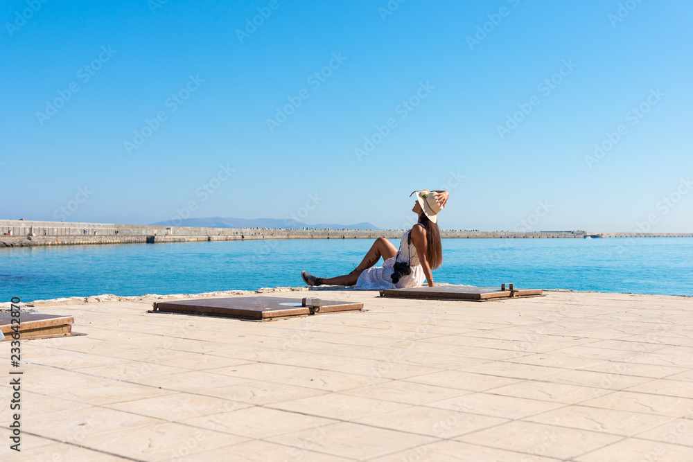 Happy tourist girl on holiday trip to Heraklion, Crete, Greece. Young woman traveler enjoyes sunshine sitting on the dock of Heraklion Venetian port marina. Dragon Island at background.