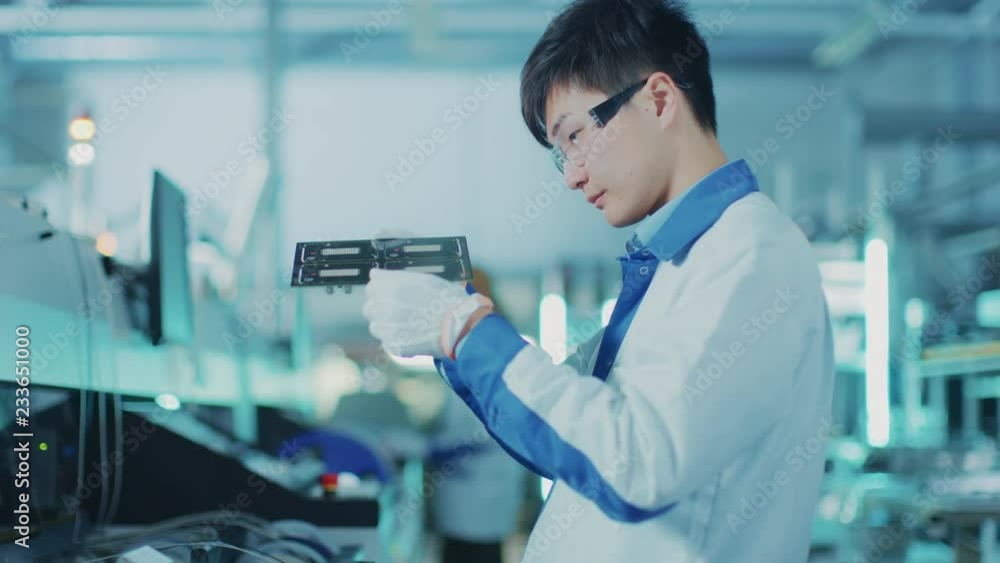 High-Tech Factory: Quality Control Engineer Checks Electronic Printed Circuit Board it for Damages. In the Background Assembly Line for PCB with Surface Mount Pick and Place Technology. High-Tech Factory: Quality Control Engineer Checks Electronic Printed Circuit Board it for Damages. In the Background Assembly Line for PCB with Surface Mount Pick and Place Technology.