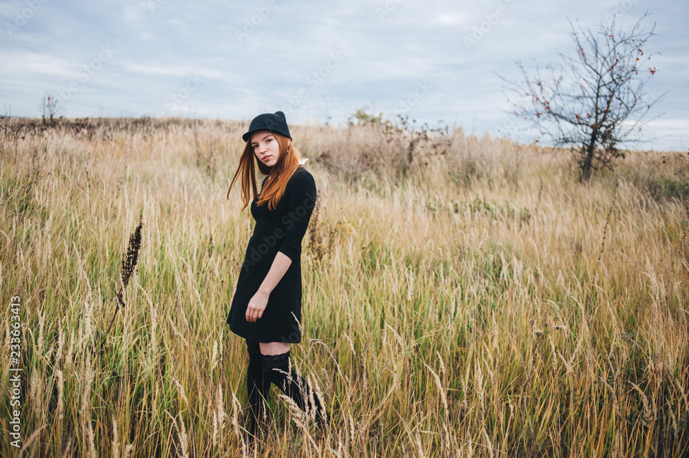 beautiful red-haired woman in a black dress walks on an autumn field.