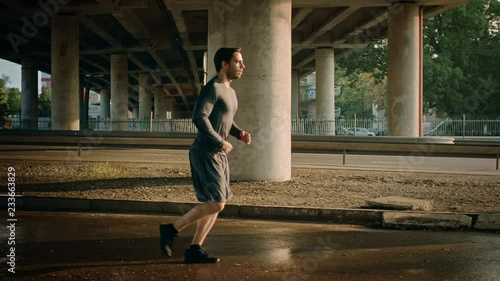 Athletic Young Man in Sports Outfit is Jogging in the Street. He is Running in an Urban Environment Under a Brindge with Cars in the Background. photo