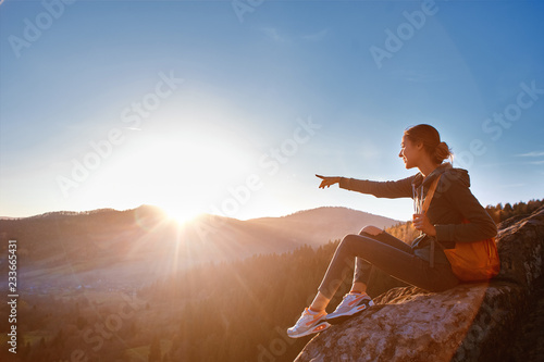 woman hiker with backpack sits on edge of cliff against background of sunrise