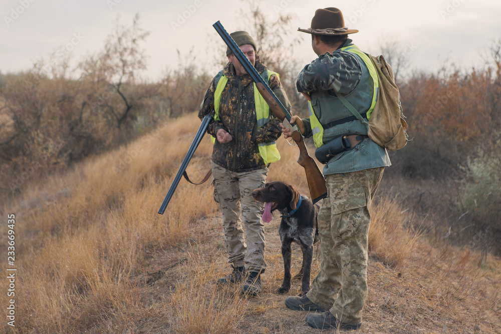 Two hunters with guns and a dog on a hill against the blue sky Two hunters with guns and a dog on a hill against the blue sky