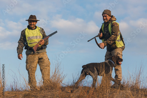 Two hunters with guns and a dog on a hill against the blue sky