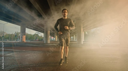 Athletic Muscular Young Man in Sports Outfit Jogging in a Street Filled With Steam. He is Running in an Urban Environment Under a Brindge with Cars in the Background. photo