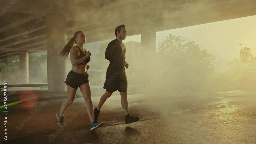 Strong and Beautiful Athletic Fitness Couple in Workout Clothes Jogging in a Street Filled With Steam on a Sunny Day. They are Running in an Urban Environment Under a Brindge with Cars Strong and Beautiful Athletic Fitness Couple in Workout Clothes Jogging in a Street Filled With Steam on a Sunny Day. They are Running in an Urban Environment Under a Brindge with Cars
