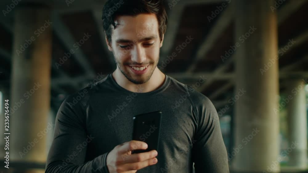 Strong Athletic Young Man in Black Sports Outfit is Using a Smartphone and Smiling on a Street. He is in an Urban Environment Under a Bridge with Cars in the Background. Strong Athletic Young Man in Black Sports Outfit is Using a Smartphone and Smiling on a Street. He is in an Urban Environment Under a Bridge with Cars in the Background.