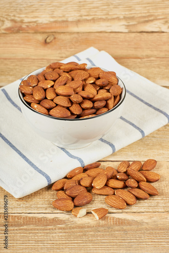 Peeled almond nuts in the white bowl and white napkin on wooden table with copy space, close-up