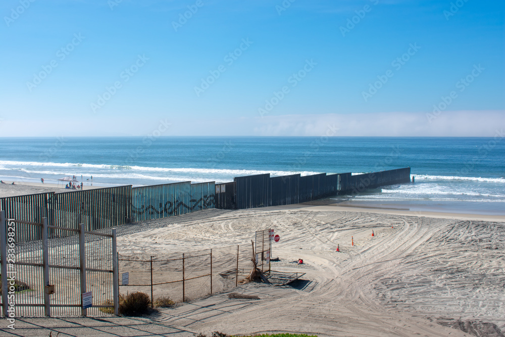 The border fence on the US - Mexico border undulates down the hills into the surf of Pacific ocean. The second border fence ends on the sandy beach. There are an empty security corridor between fences